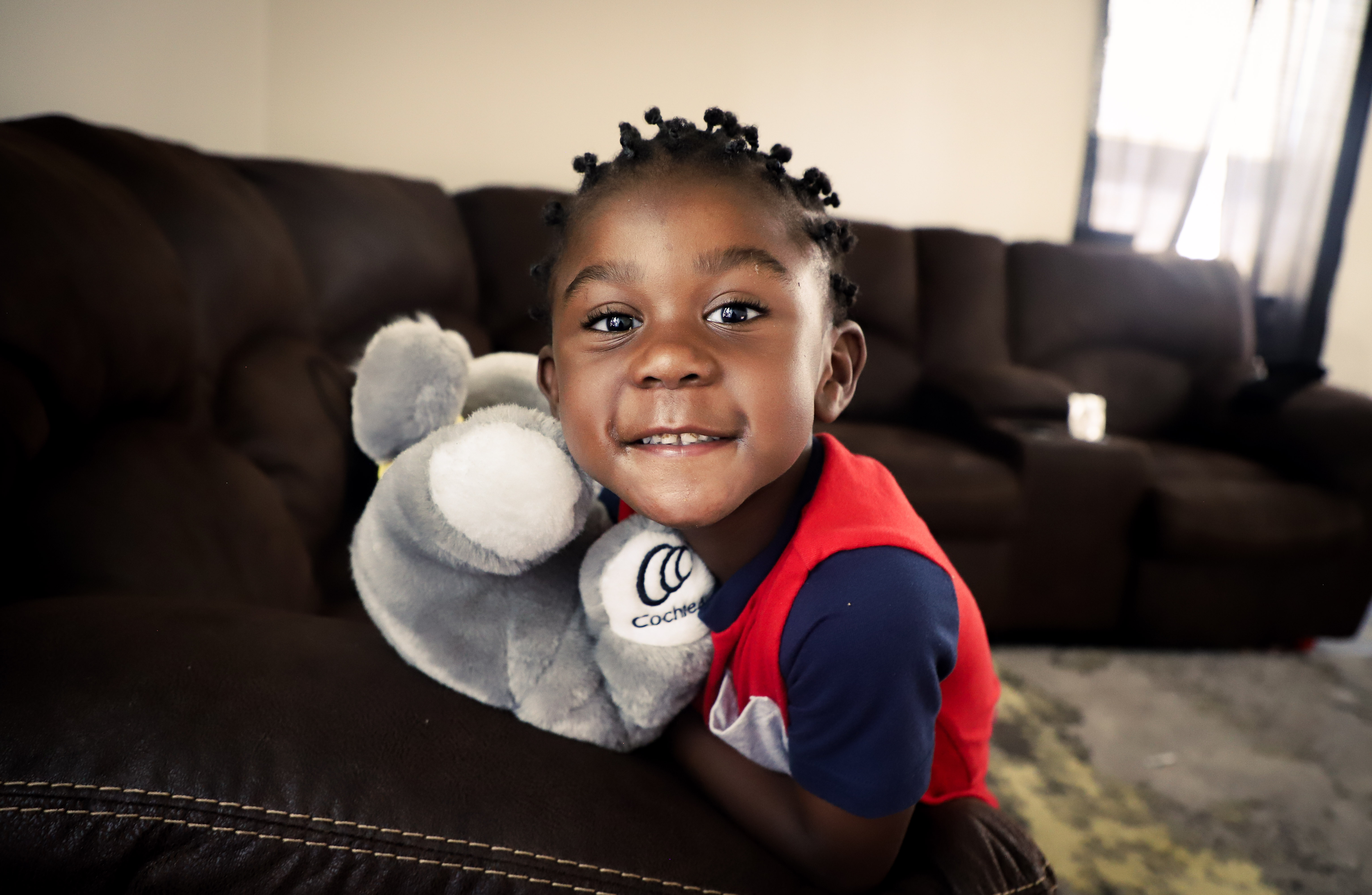 Child leaning against a brown couch with a grey stuffed animal