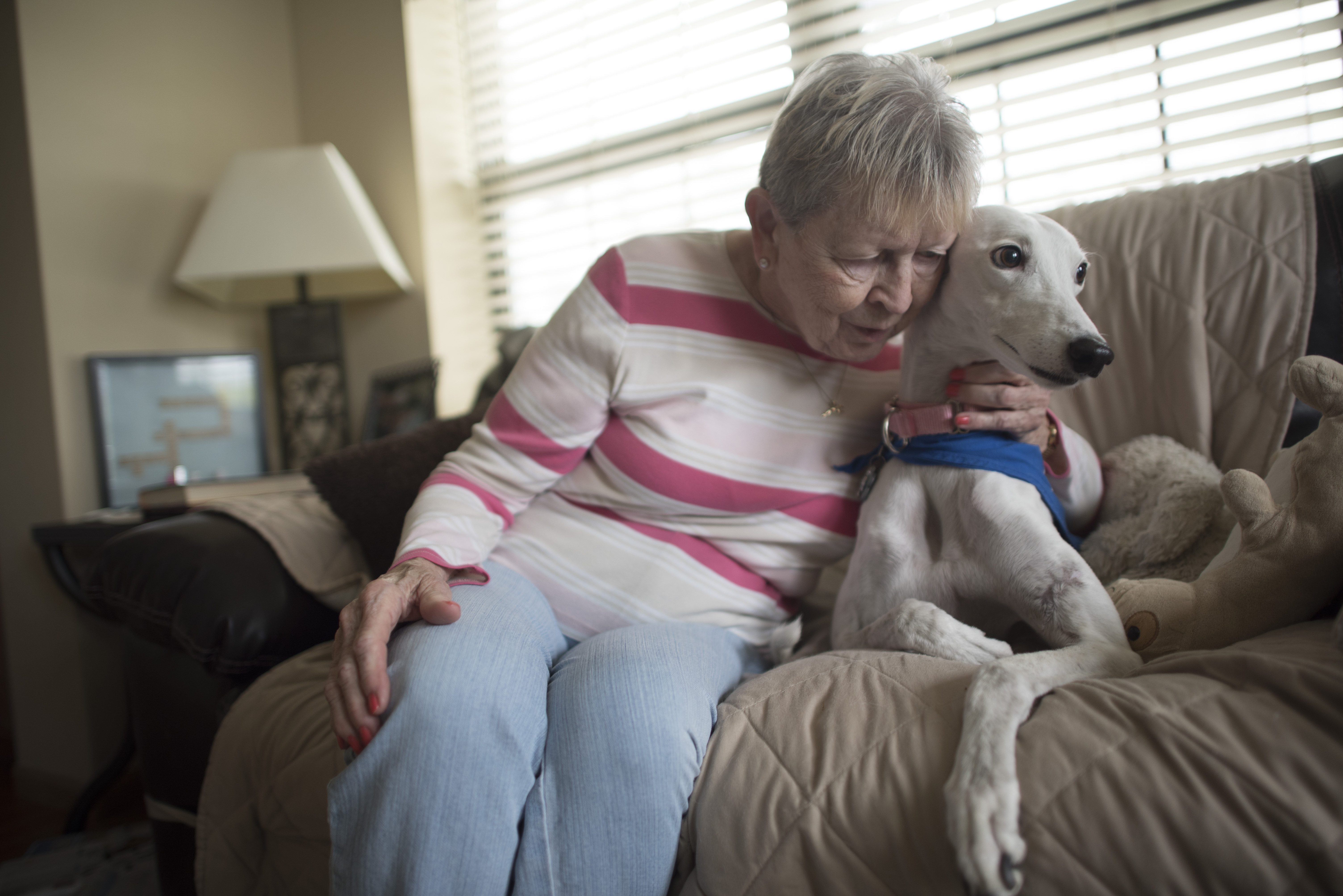Older woman sits on a couch and hugs a white dog with one arm. There's a side table, lamp, and a window with blinds partially closed in the background.